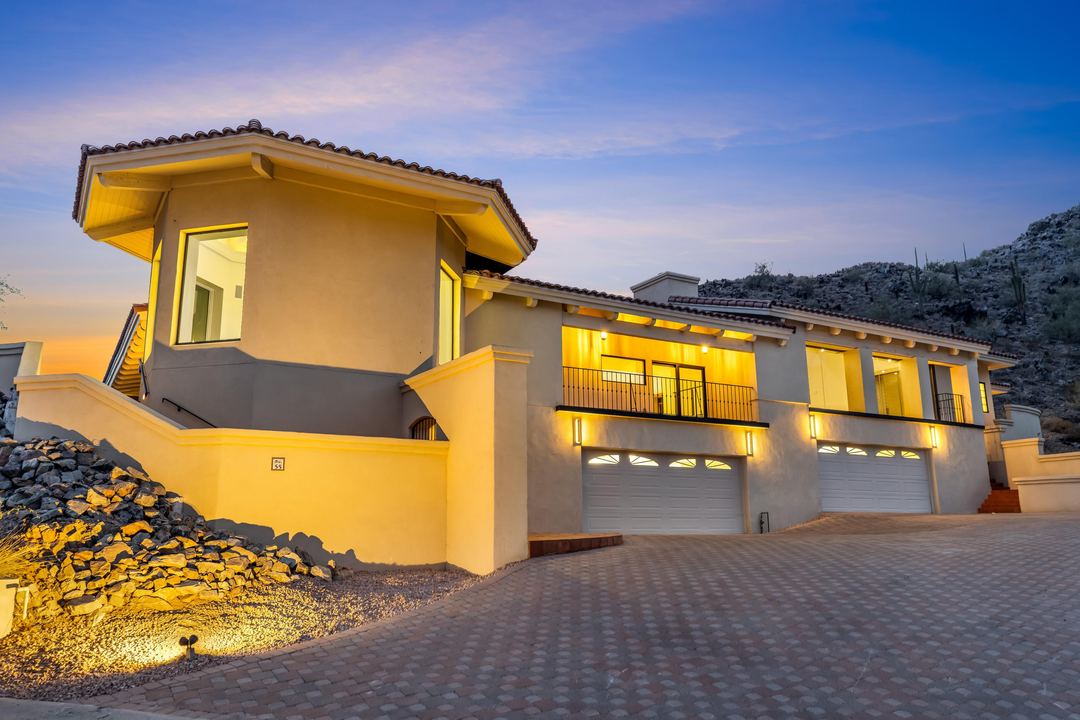 Two-story hillside home at dusk with uplit stone and three lit garage doors