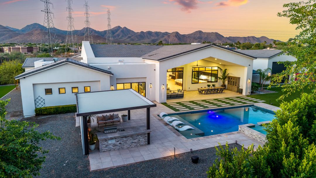 Twilight aerial over a backyard pool, outdoor kitchen and turf lawn, with Camelback Mountain on the horizon