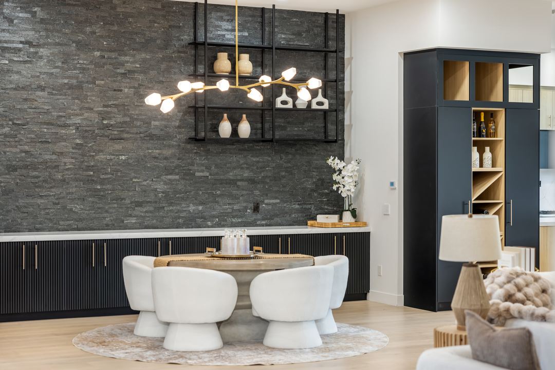 Dining table and white chairs under a sculptural chandelier against a dark stacked-stone wall