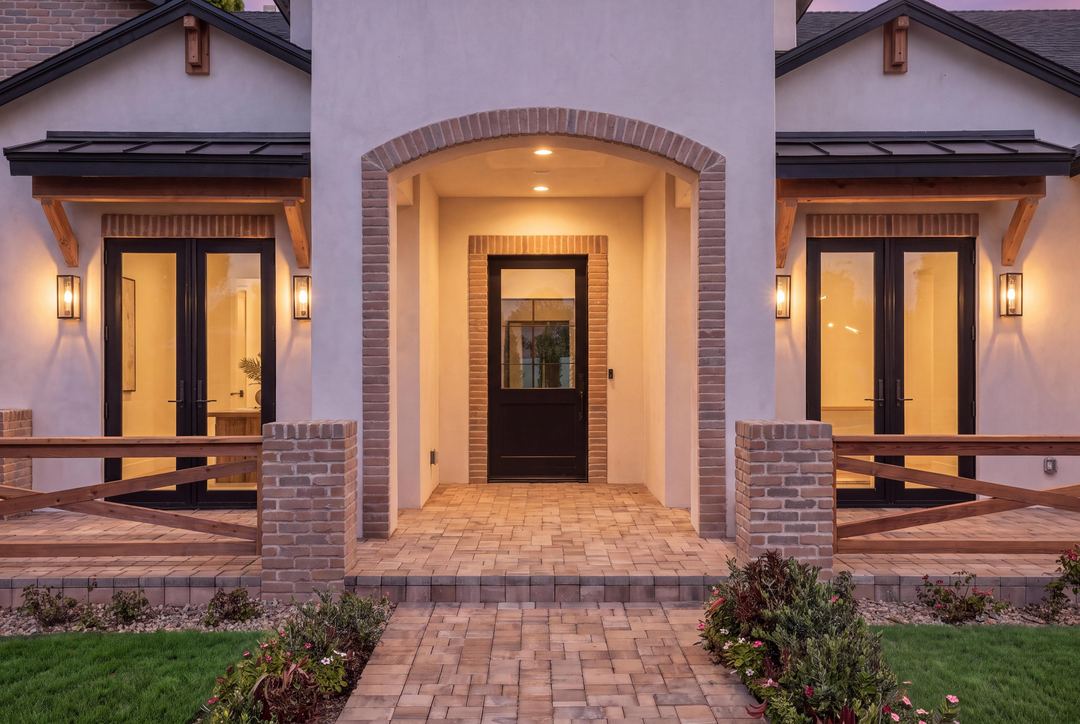 Brick-arched front entry at dusk, a black steel door between lit lanterns and wood rail fencing on the porch