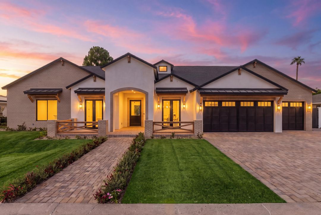 Modern farmhouse at dusk with a brick-arched entry, lit windows and a three-car garage, a paver drive beside the lawn under a pink sky