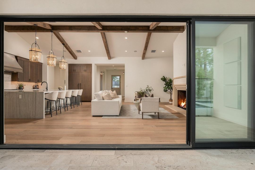 The great room seen from the patio through open sliding glass doors, with a beamed ceiling, kitchen pendants, a cream sectional and the fire lit