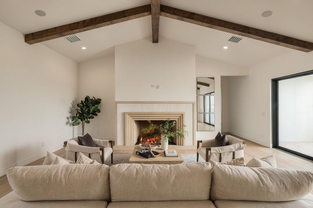 Great room with a vaulted beamed ceiling, a cream sectional, two armchairs and a fluted stone fireplace with the fire on