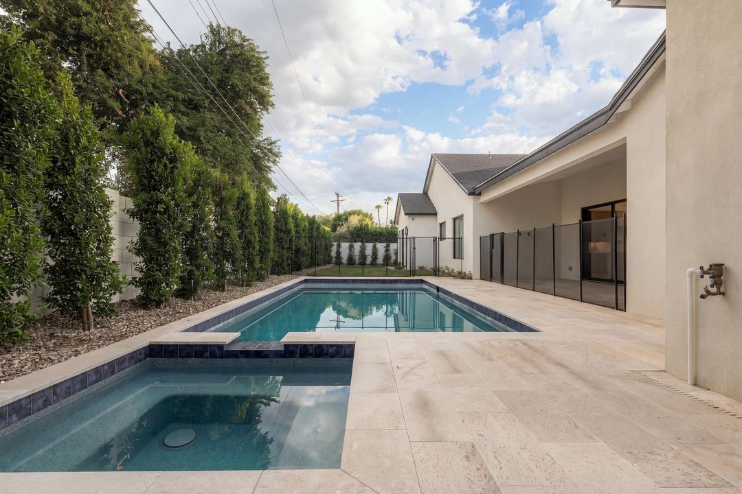 Pool and spa in daylight on a travertine deck, a hedge of trees along the wall and the house beyond