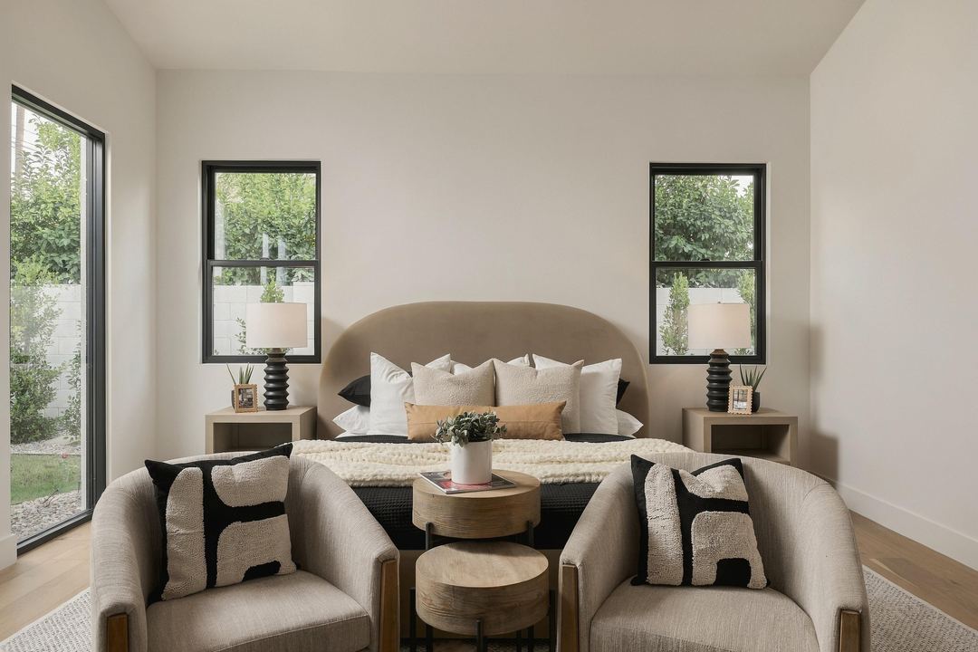 Tan upholstered bed with layered pillows between two black-framed windows, two armchairs and nesting tables at its foot